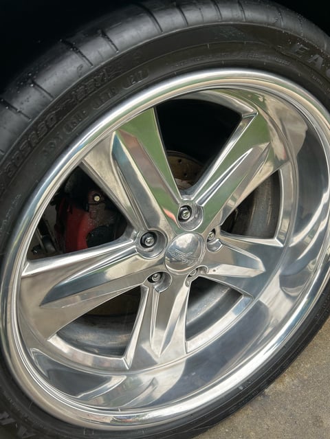 Close-up of a polished chrome custom car wheel with red brake calipers visible through the spokes and a black tire mounted on top